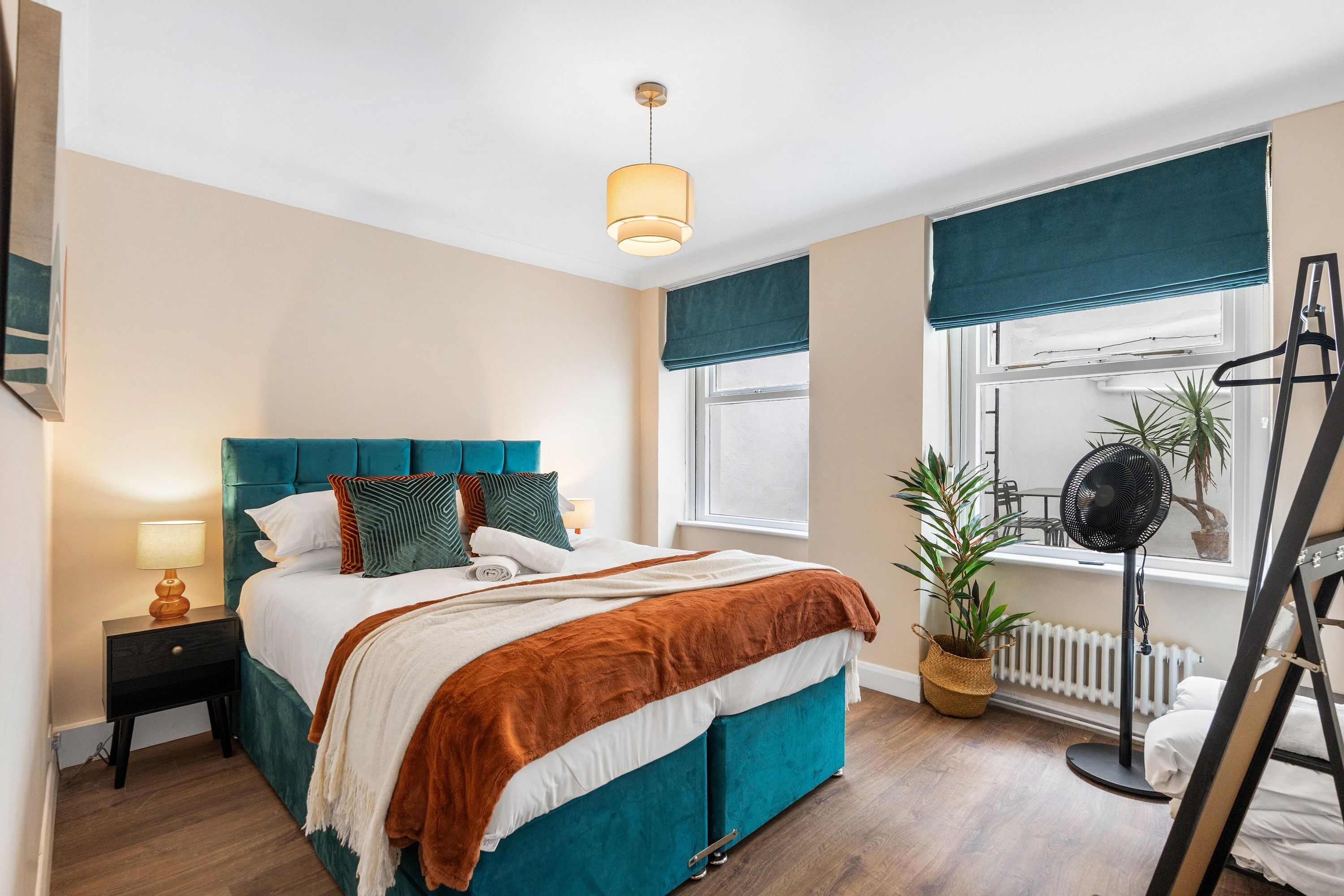 A bedroom with a bed, bedside table, lamp, window with curtains, radiator, and a potted plant.