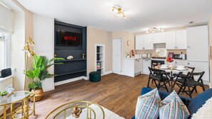 A modern living room with a flat-screen TV, a dining area with a table and chairs, and a kitchen with white cabinets. - Coastal Calm By My Getaways (Brighton)