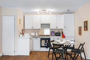 A compact kitchen with white cabinets, a dishwasher, a stove, and a dining area with a table and chairs. - Coastal Calm By My Getaways (Brighton)