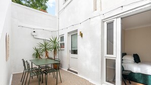 A small patio with a table and chairs, a potted plant, and a sliding glass door leading to a bedroom with a bed and pillows. - Coastal Calm By My Getaways (Brighton)