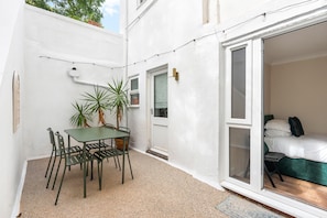 A small patio with a table and chairs, a potted plant, and a sliding glass door leading to a bedroom with a bed and pillows. - Coastal Calm By My Getaways (Brighton)