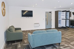 Ein modernes Wohnzimmer mit einer Couch, einem an der Wand montierten Fernseher und einer Küchenzeile mit offenem Kühlschrank.