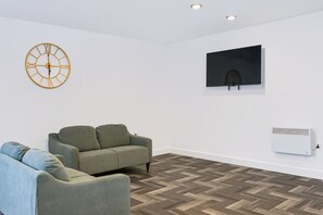 A room with a grey sofa, a wall-mounted TV, a decorative clock, and a white air conditioning unit. - Brand New Spacious Studio in Beeston (Beeston)
