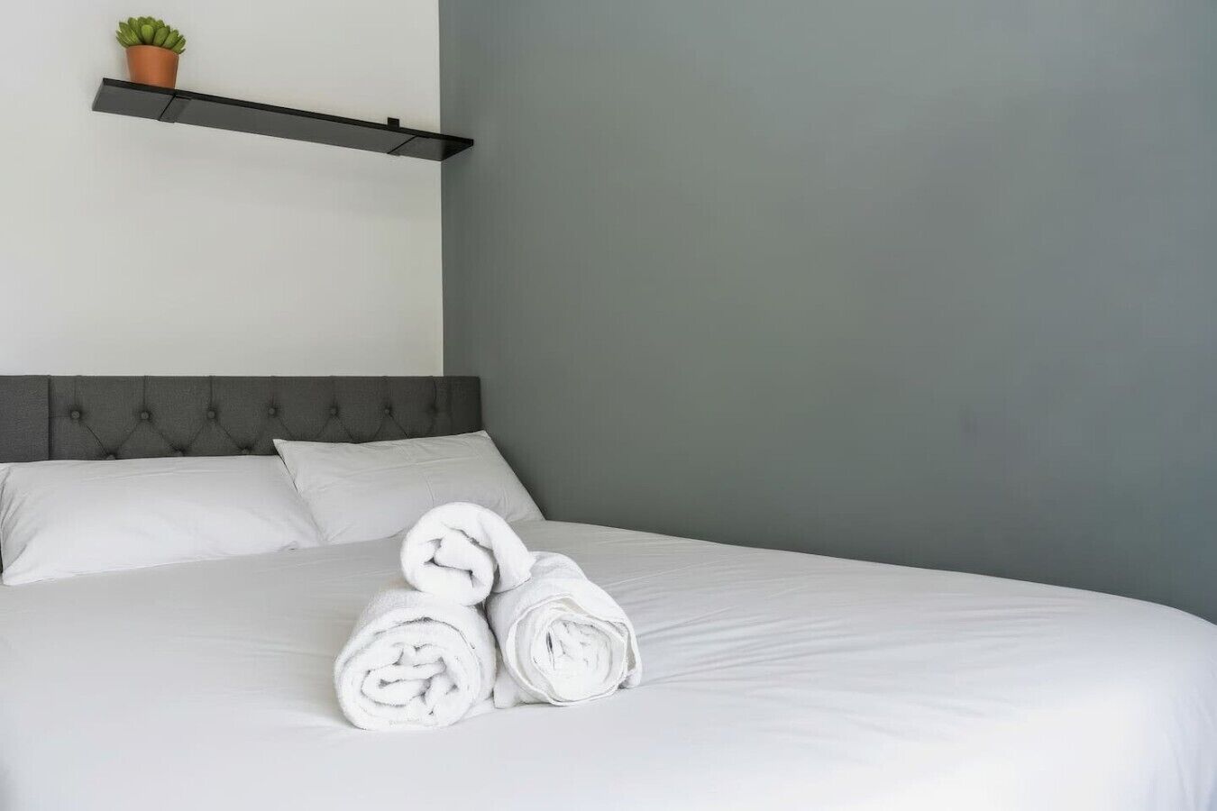 A neatly made bed with white linens and rolled towels, a grey headboard, and a small potted plant on a shelf above the bed.