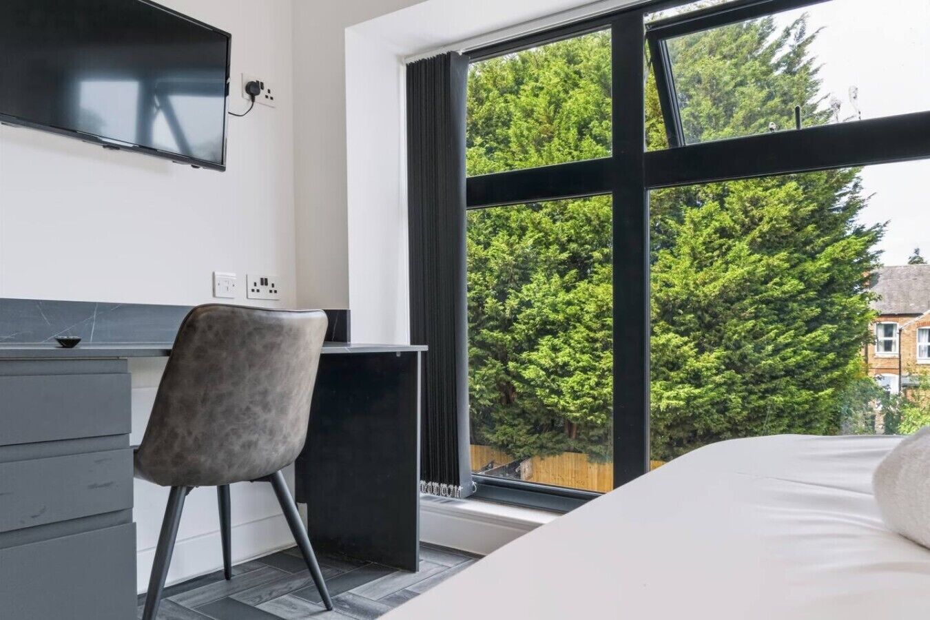 A bedroom with a bed, a desk, and a chair. There is a window with a view of trees.