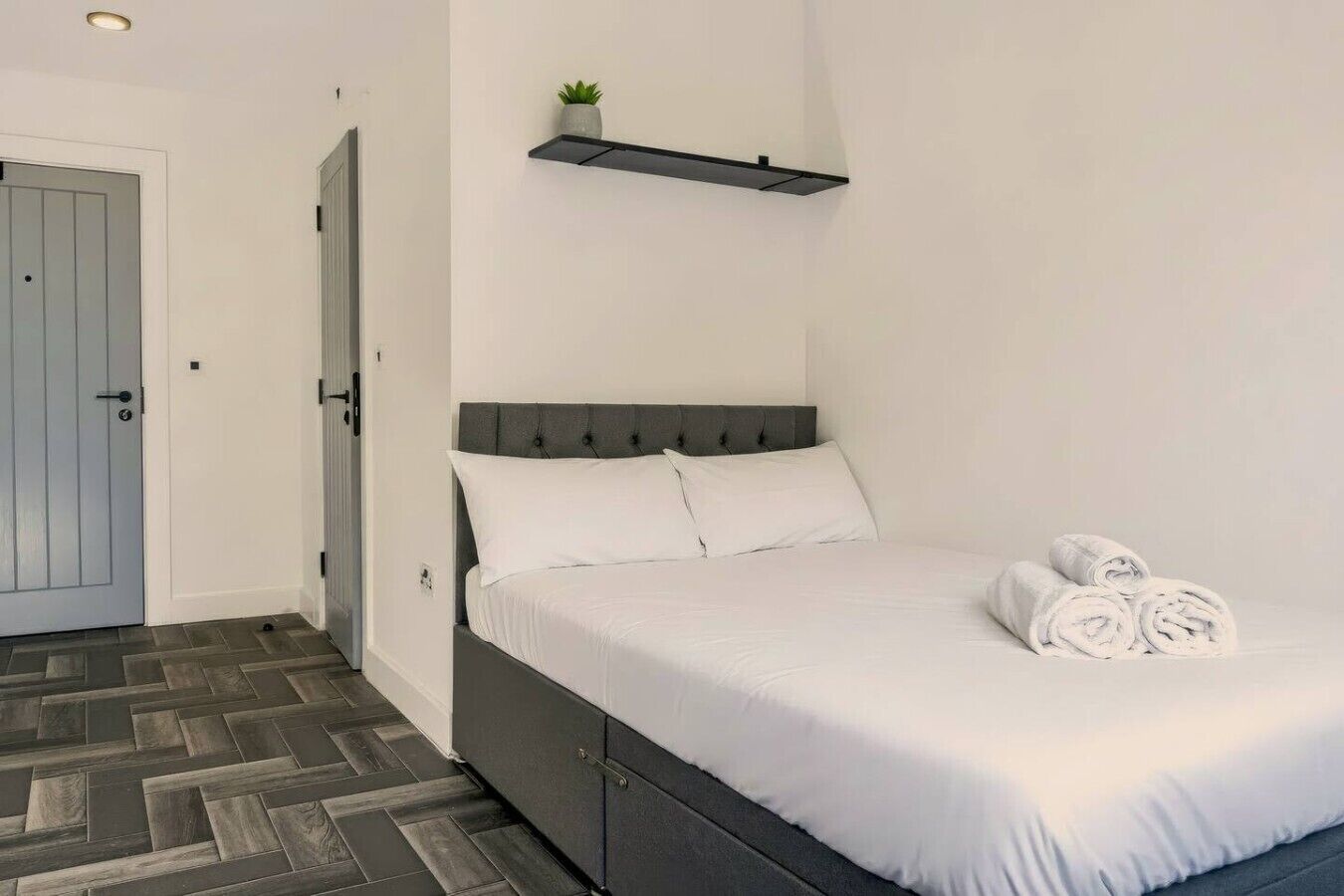 A neatly made bed with white linens and rolled towels, a grey headboard, and a small plant on a shelf above.