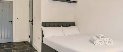A neatly made bed with white linens and rolled towels, a grey headboard, and a small plant on a shelf above.