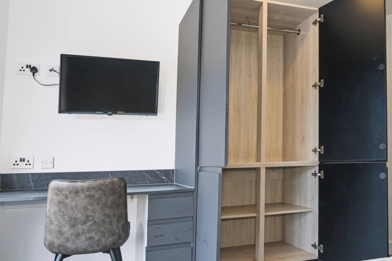 Una habitación con televisión de pantalla plana montada en la pared, una silla gris y un armario con varios compartimentos.