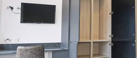 A room with a flat-screen TV mounted on the wall, a grey chair, and a wardrobe with multiple compartments.