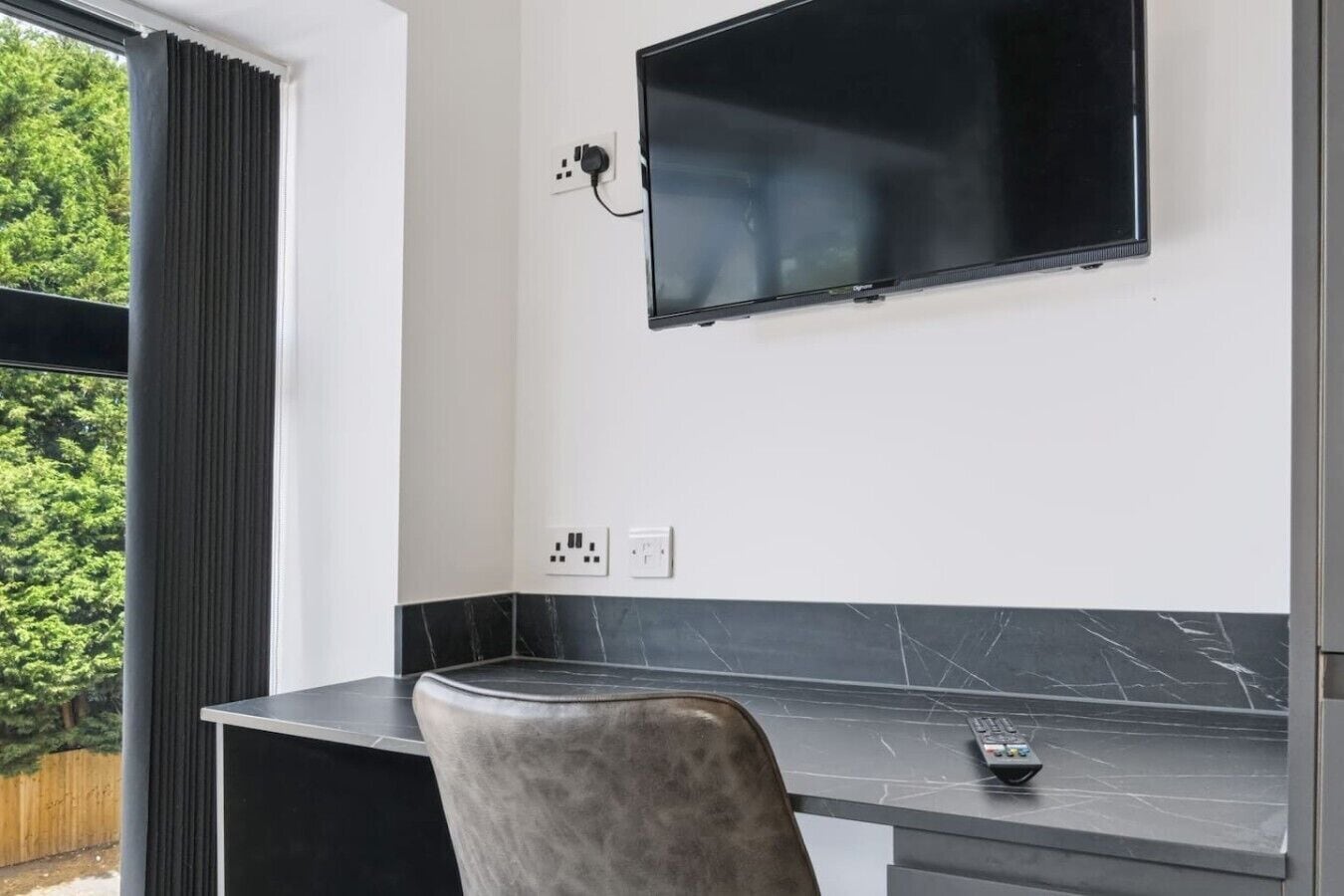 Quarto moderno com televisão de tela plana na parede, escrivaninha com controle remoto e uma cadeira.