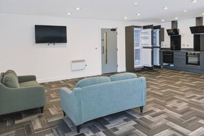 A modern living room with a flat-screen TV, a sofa, and a kitchen area with built-in appliances. - Brand New Spacious Studio in Beeston (Beeston)