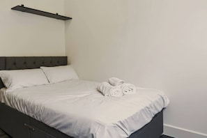 A bed with a grey headboard, white bedding, and rolled towels on the side. - Brand New Spacious Studio in Beeston (Beeston)