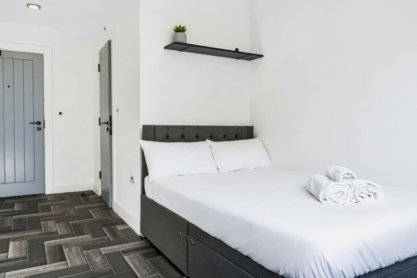 A bedroom with a bed, a door, a wall-mounted shelf with a plant, and a herringbone-patterned floor.