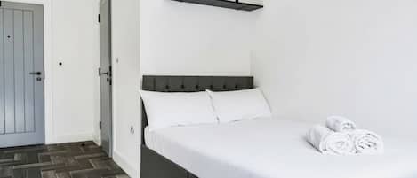 A bedroom with a bed, a door, a wall-mounted shelf with a plant, and a herringbone-patterned floor.