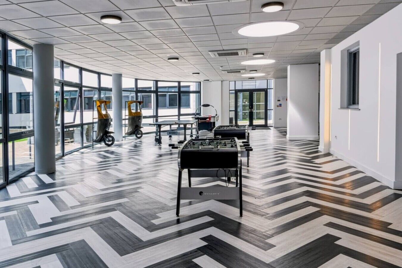 Un vaste espace intérieur doté d’un sol à motifs, de grandes fenêtres et d’une table de baby-foot.