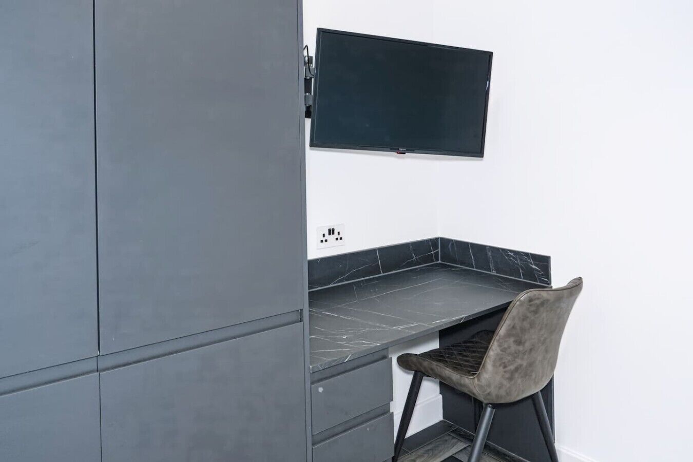 Une petite cuisine équipée d’une cuisinière intégrée, d’un téléviseur à écran plat fixé au-dessus du comptoir, et d  ’une chaise dotée d’un siège et d’un dossier rembourrés.