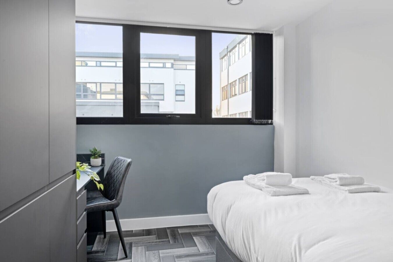 A hotel room with a bed, a desk, a chair, and a window with a view of buildings.