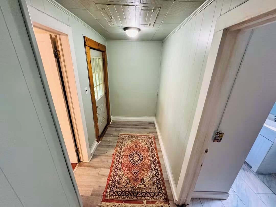 A narrow hallway with a patterned rug, a wooden door, and a white wall.