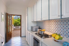 Une cuisine avec des meubles blancs, une cuisinière à gaz, un grille-pain et une vue sur la verdure par une porte ouverte.