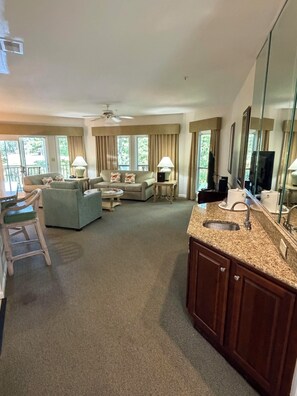 A hotel room with a living area, a sofa set, a dining table, a kitchen area with a sink, and large windows.