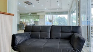 A black leather sofa in a modern room with a large window, a clock on the wall, and a ceiling with recessed lighting. - Premium Family Suite With Balcony Mauji's (Prayagraj)