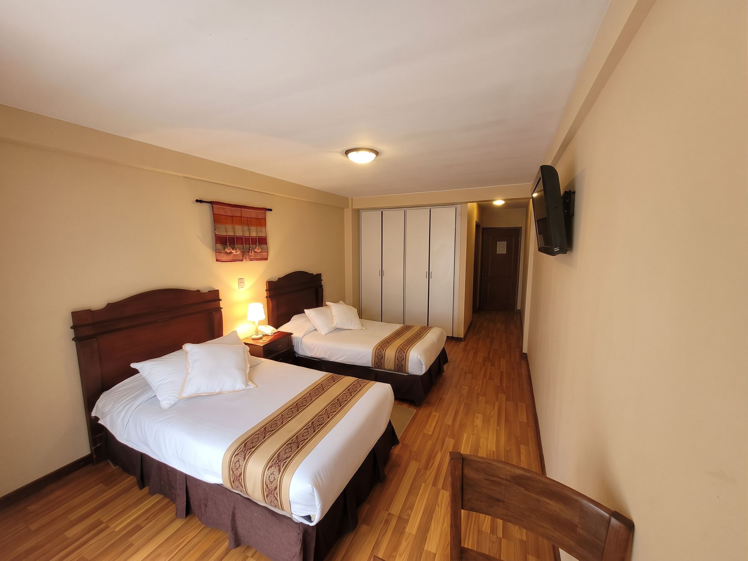 Habitación de hotel con dos camas, piso de madera, armario y televisor montado en la pared.
