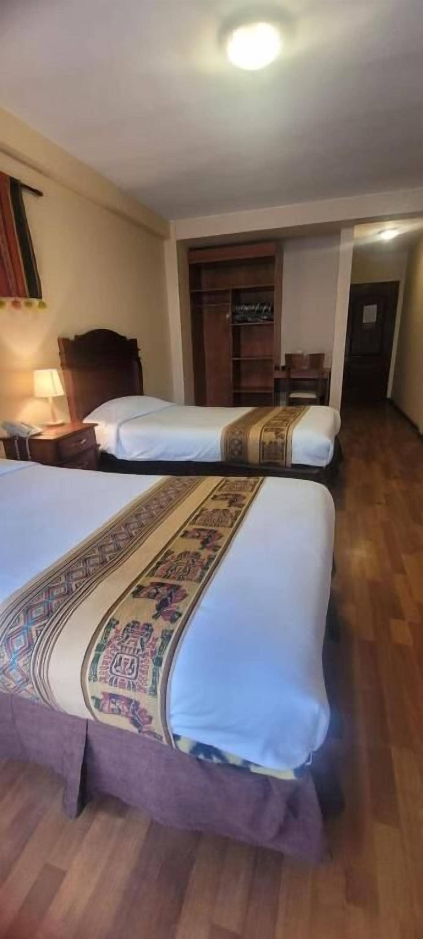 Quarto de hotel com duas camas, piso de madeira e mesa de cabeceira com abajur.