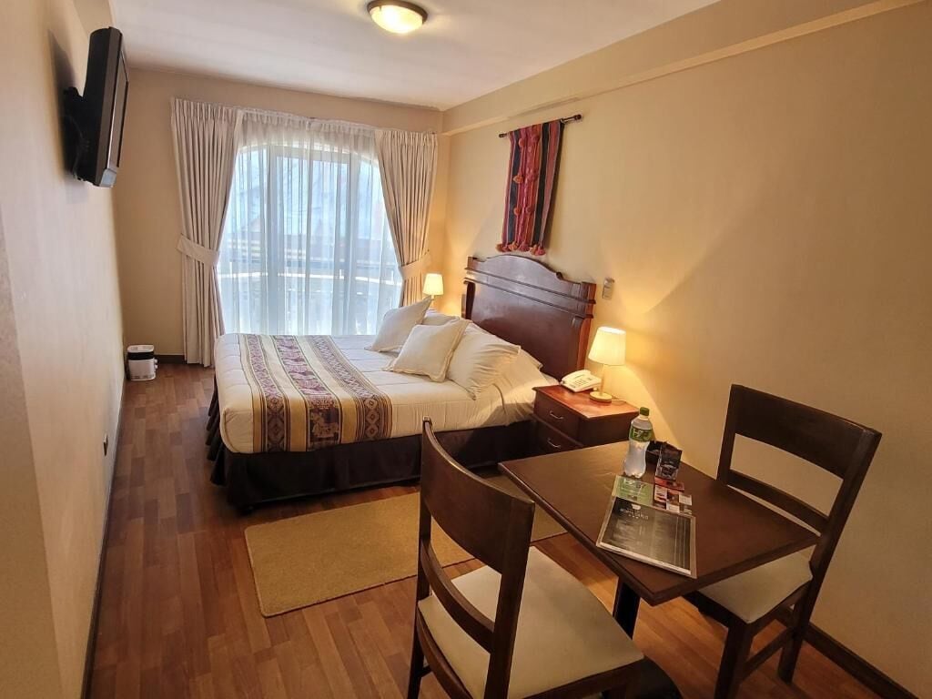Habitación de hotel con cama, mesita de noche, lámpara y una pequeña mesa con sillas.