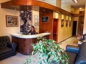 Una zona de recepción con un mostrador de madera, una recepcionista y una planta grande en maceta.