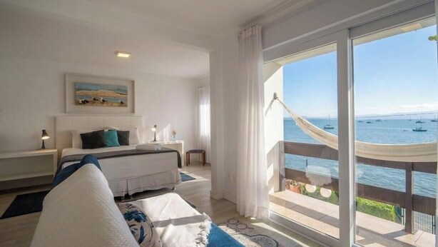 A hotel room with a bed, a bedside table, a nightstand, and a large window with a view of the sea and boats. - Beautiful Beachfront House Santo Antonio Cda1647 (Florianópolis)