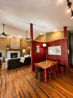 A room with a stone fireplace, a wooden dining table with chairs, a sofa, and a ceiling fan. - 5-bedroom lakefront home with private dock, swim platform, fishing & more! (Camdenton)
