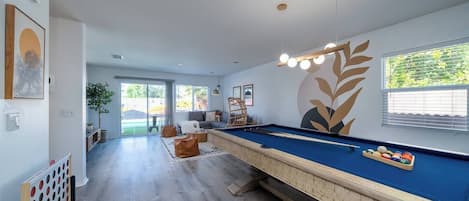 Una mesa de billar en una habitación con un ventanal grande, una planta y un muro con diseño de hojas.