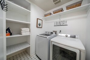 A laundry room with a washing machine, dryer, and shelves with folded towels. - private pool + spa + game room + near Coachella | Family & Group Friendly (Indio)