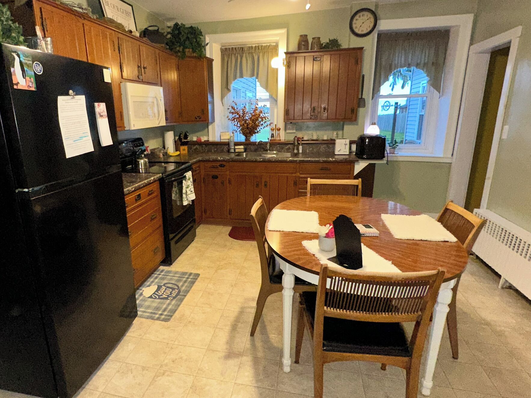 Una cocina con armarios de madera, un refrigerador negro, una mesa de comedor con sillas y una ventana con cortinas.