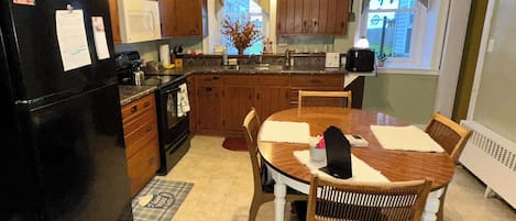 Una cocina con armarios de madera, un refrigerador negro, una mesa de comedor con sillas y una ventana con cortinas.