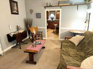 A living room with a wooden table, a sofa, a dining area, and a kitchen visible through an open doorway.