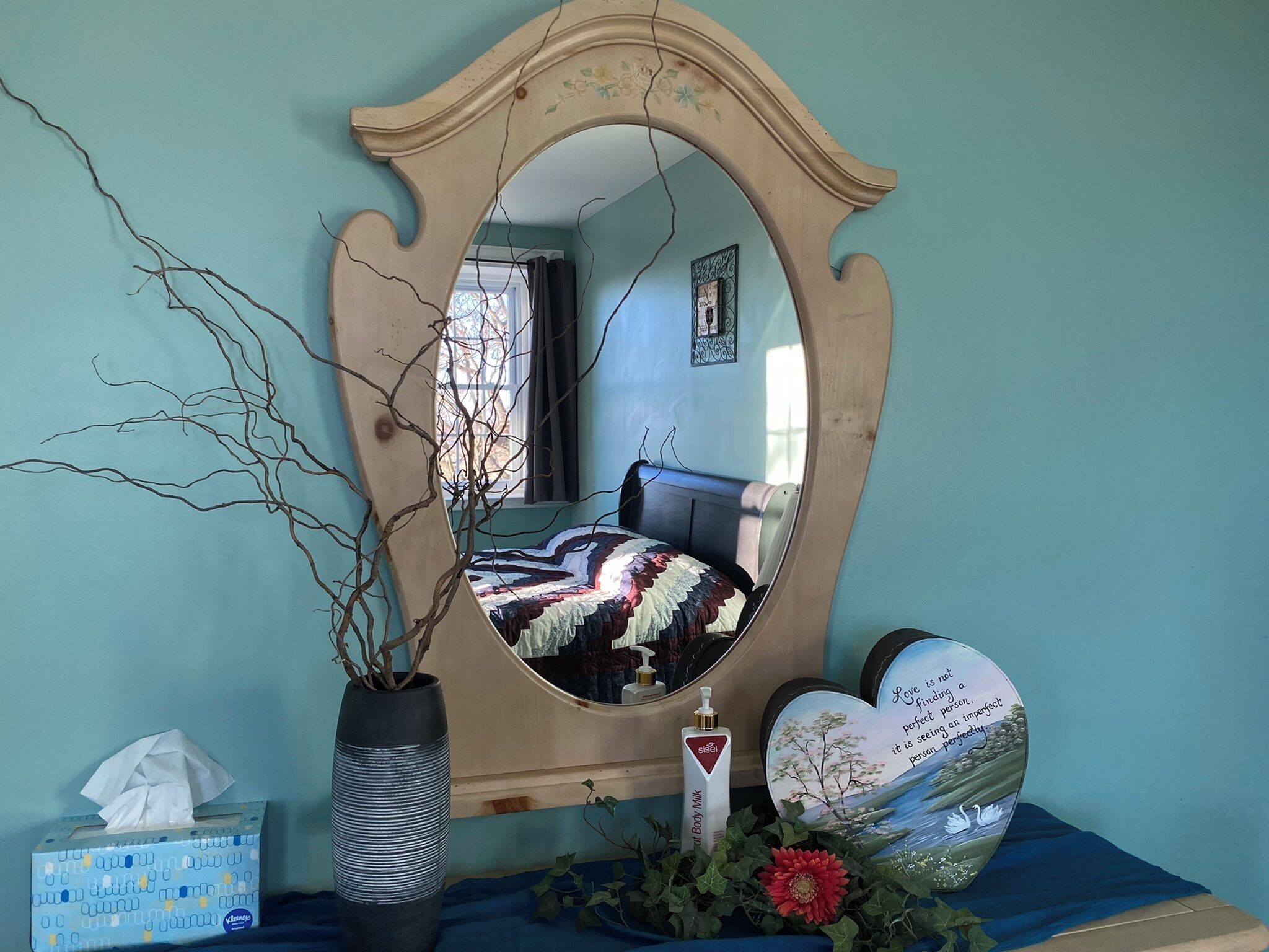 Une chambre à coucher comprenant un lit, un miroir, un vase avec des branches, une boîte de mouchoirs et des objets décoratifs.
