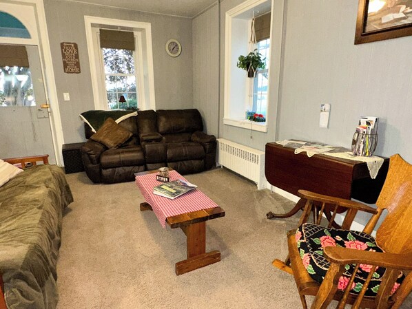 A cozy living room with a brown leather sofa, a wooden coffee table with a checkered tablecloth, a wooden chair with floral upholstery, and a window with a view of trees.