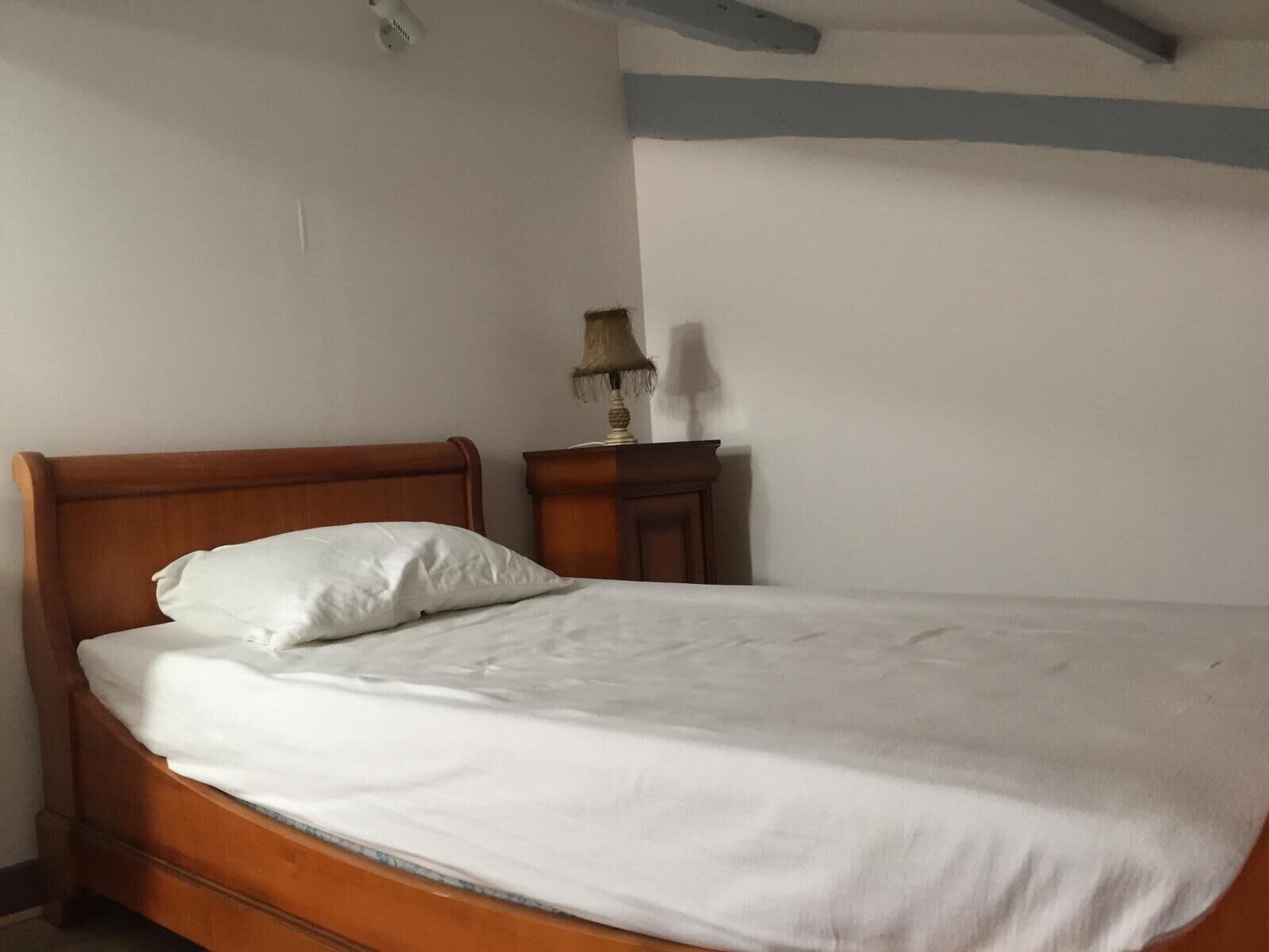 Ein Einzelbett mit Holz-Kopfteil und weißer Bettwäsche in einem Raum mit schlichten Wänden und einem kleinen Nachttisch mit Lampe.