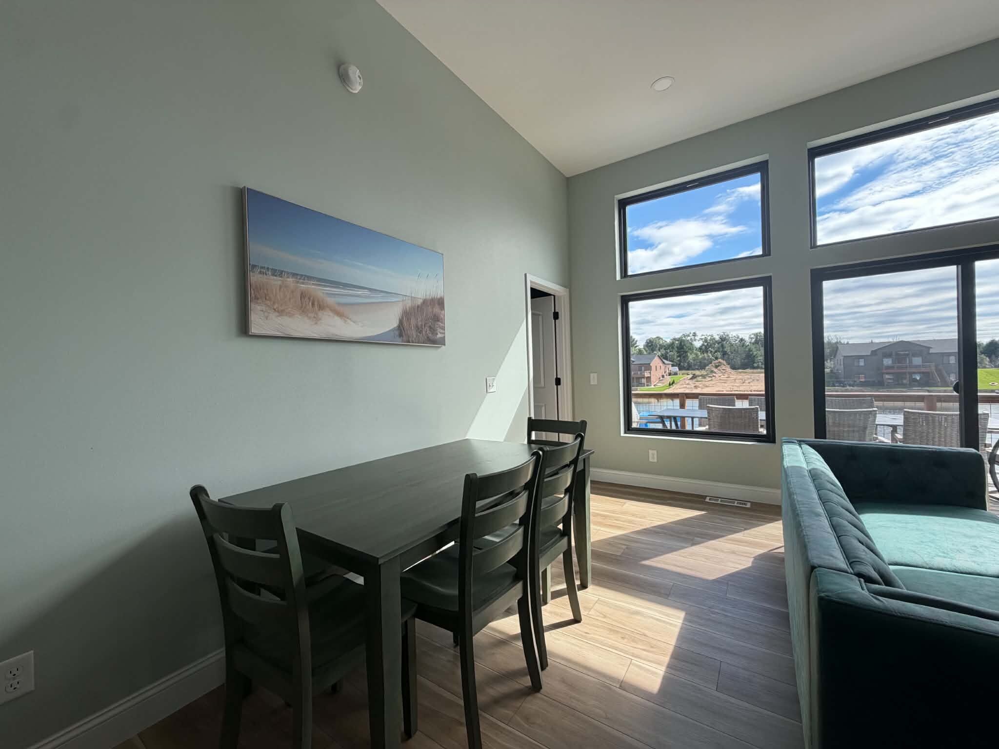 Comedor con mesa y sillas, un cuadro en la pared y ventanas amplias con vistas al exterior.