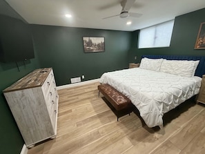 Ein Schlafzimmer mit Bett, einer Bank, einem Schminktisch, einem Deckenventilator und einem Fenster mit Jalousien.