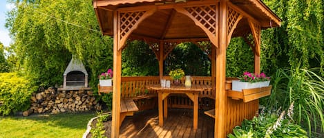 A wooden gazebo with a table and benches, surrounded by lush greenery and a stone fireplace.