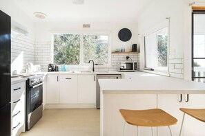 A modern kitchen with white cabinetry, a black refrigerator, a microwave, and a sink. There are two wooden stools and a window providing natural light. - Bayview Cottage Serene Escape between Beach and Bay (Blairgowrie)