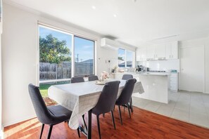 A modern kitchen with white cabinets, a dining area with a table and chairs, and a view of a backyard through large windows. - Coastal Grove—Sunlit Stay near Beach & Hot Springs (Tootgarook)