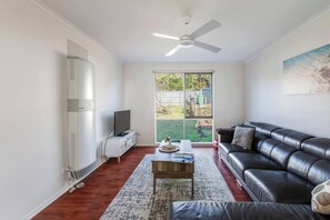A modern living room with a flat-screen TV, a leather sofa, a coffee table, and a ceiling fan. - Coastal Grove—Sunlit Stay near Beach & Hot Springs (Tootgarook)