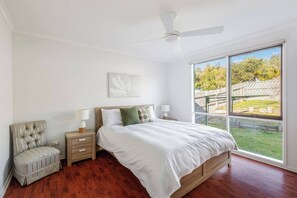 A bedroom with a bed, bedside tables, a chair, a ceiling fan, and a large window with a view of a backyard. - Coastal Grove—Sunlit Stay near Beach & Hot Springs (Tootgarook)