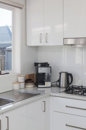 A modern kitchen with white cabinets, a coffee maker, a kettle, and a window providing a view of the outside. - Coastal Grove—Sunlit Stay near Beach & Hot Springs (Tootgarook)