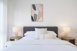 Una cama bien hecha con sábanas blancas y varios almohadones, con dos lámparas de noche sobre mesitas de madera redondas.