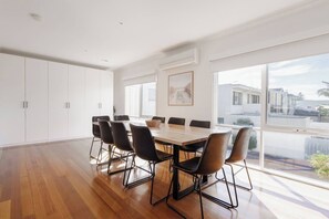 A spacious dining area with a wooden table and black chairs, large windows with a view of buildings, and white cabinets. - Sea Quinn Escape Steps to Dromana Beach and Dining (Dromana)