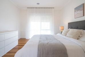 Ein Schlafzimmer mit einem großen Bett, einem Nachttisch mit Lampe und einem Fenster mit durchsichtigen Vorhängen.